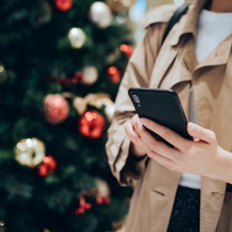 apgriezts kadrs un sievietes vidusdaļa, kas izmanto viedtālruni krāsainas Ziemassvētku eglītes priekšā svētku Ziemassvētku sezonā