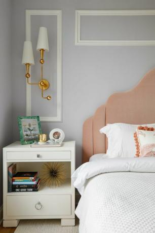 guļamistaba, gulta ar baltu linonu, balts gultas galds, grāmatas, rozā galvas klājs, baltas un zelta krāsas skapīši, balta vainaga veidne uz gaiši pelēki krāsotām sienām