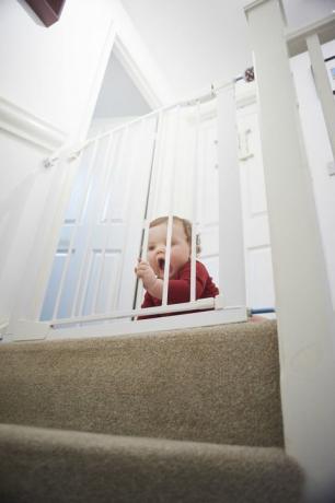 Mazuļa drošības vārti uz kāpnēm: Zēns uz savas mājas laukuma. Viņš atrodas aiz mazuļa drošības vārtiem, kas viņu attur no kāpņu telpas briesmām.