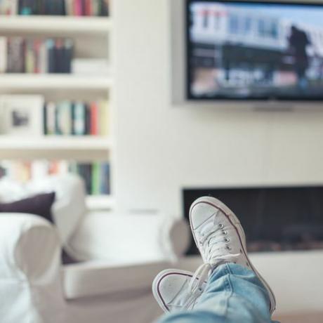 Džinsu un čības attēls, kājas uz dīvāna, ar televizoru, kamīnu, dīvānu un grāmatu skapi fonā.